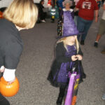 A child dressed as a witch in a purple costume holds a treat bag while people walk around during a Halloween event.
