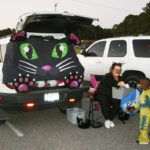 A decorated car trunk looks like a black cat; a woman in cat ears gives candy to a child in a costume.