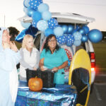 Three women stand by a decorated car trunk with blue balloons, a pumpkin, and a yellow surfboard, participating in a festive outdoor event.