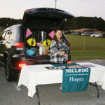 Woman stands behind a table with informational materials, in front of a car trunk decorated with large cat eyes and ears. Banner reads "McLeod Hospice."