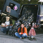 Two people in costumes sit by a decorated car trunk with Halloween props, including skeletons and spooky figures, during a trunk-or-treat event outdoors.