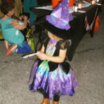 Young child in a purple witch costume and hat holding paper, standing on a street during a Halloween event.