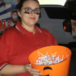 Woman in red shirt holding an orange bucket filled with lollipops, standing outdoors, with a decorated car and signs in the background.