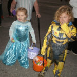 Two young children in Halloween costumes, one dressed as a princess and the other as a robot or superhero, holding candy buckets on a street.