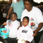 A woman with two children at an outdoor event, one child holds a blue bucket, all dressed casually with people in the background.