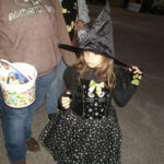 A young girl dressed as a witch in a black hat and dress stands next to an adult holding a Halloween candy bucket.