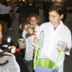 Two children in white lab coats trick-or-treating at night, holding bags and stuffed animals, with other people in the background.