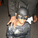 A young child dressed in a Batman costume, including a black mask and suit, stands in front of an adult whose hand rests on the child's shoulder.