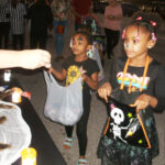 Two young girls in costumes holding bags, participating in trick-or-treating at a Halloween event, with people and decorations visible in the background.