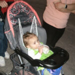 A baby in a stroller wearing fairy wings drinks from a bottle while adults stand nearby.