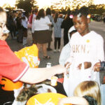 Children in costumes receive candy from an adult at an outdoor event, with more people gathered in the background during daylight.