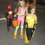 Three children in colorful Halloween costumes walk together, carrying treat bags and buckets, smiling and enjoying trick-or-treating at night.