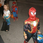 Children in costumes trick-or-treating at night, with two dressed as Spider-Man and one as a princess, holding candy buckets.