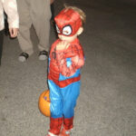 A young child dressed in a Spider-Man costume stands on pavement, holding a pumpkin-shaped container, likely during Halloween.