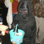 Person in a black hooded costume with red eye details holding a blue Halloween bucket filled with treats.