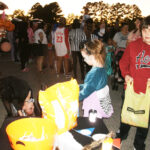Children collecting candy from buckets at an outdoor event, with a crowd of people and trees visible in the background during sunset.