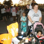 A woman pushes a stroller with a small child, accompanied by another child in costume, and people lining up outdoors near orange buckets and a table.