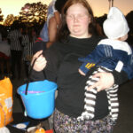 A woman holding a baby and a blue bucket stands outdoors during an evening event, surrounded by people and Halloween decorations.