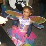 A young girl dressed as a unicorn fairy with rainbow wings and a horn, holding a pink bag, receives candy from someone at an event.