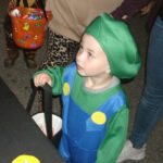 Young child in a green and blue costume with a green hat, holding a Halloween bucket, looking up indoors, surrounded by other people.