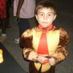 A young boy wearing a monkey costume with a red tie stands outdoors at night, holding something in his hands.
