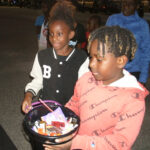 Two children holding a bucket filled with candy, standing outdoors at night, surrounded by other people.