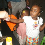 Two young girls reach for candy at a table, one holding an orange bag, both with stickers on their faces and shirts.