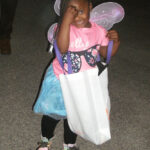 Little girl in a fairy costume with wings, holding a large white bag, standing indoors on a grey floor.