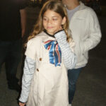 A young girl with long hair, wearing a light coat with a ribbon and striped sleeves, stands outside at night with people around her.