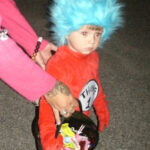A child dressed as Thing 1 from Dr. Seuss, wearing a blue wig and red costume, holds a Halloween bucket filled with candy.