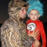Man in camouflage hoodie holding a child dressed as Thing 2 from Dr. Seuss, with a blue wig and red costume, outdoors at dusk.