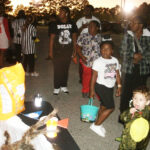 Children and adults in costumes holding baskets, gathered outdoors at dusk, possibly for a Halloween or trick-or-treat event.
