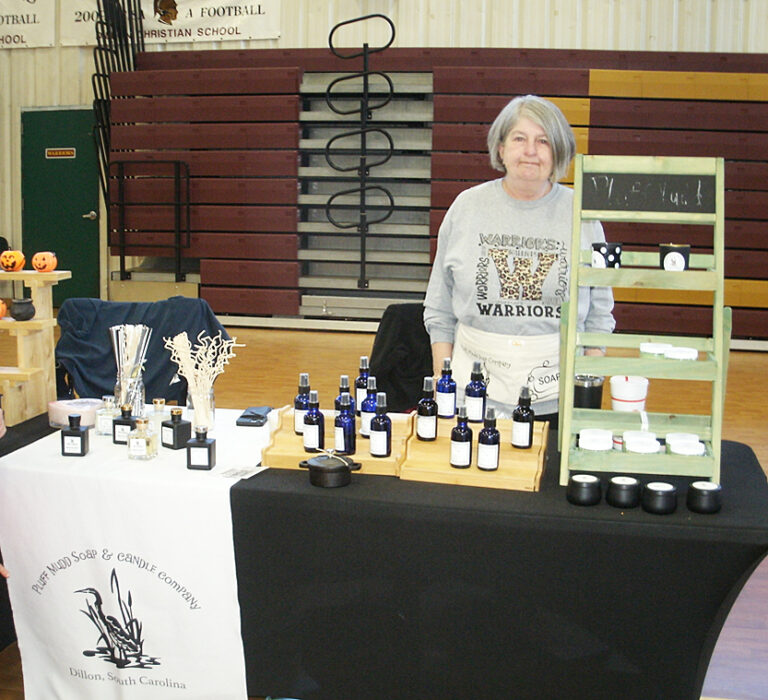 A woman stands behind a display table with bottles, jars, and products at an indoor market or fair, featuring informational materials and a banner.