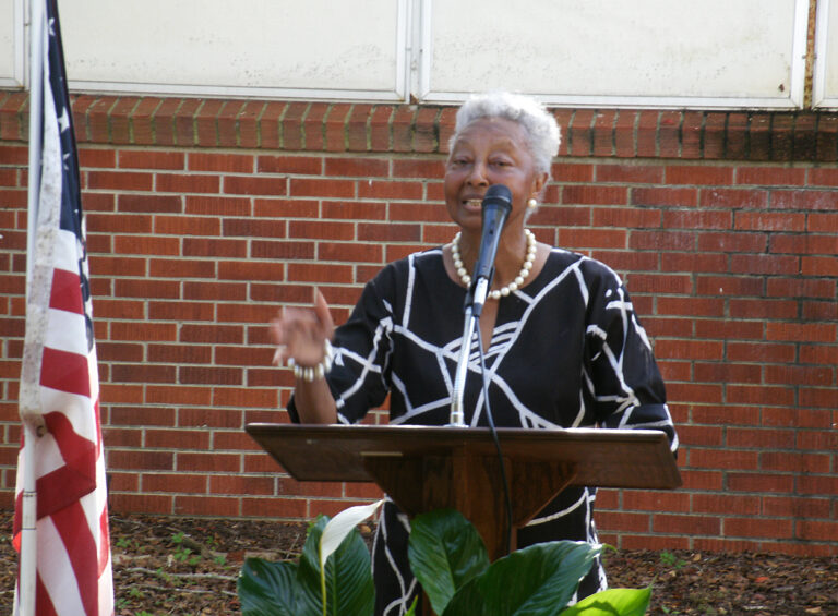 elderly woman speaks at a podium outdoors, wearing a black and white dress, with a brick wall in the background and plants in the foreground.
