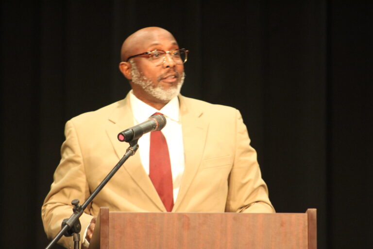 A man in a beige suit and red tie speaks at a podium with a microphone, set against a dark background.