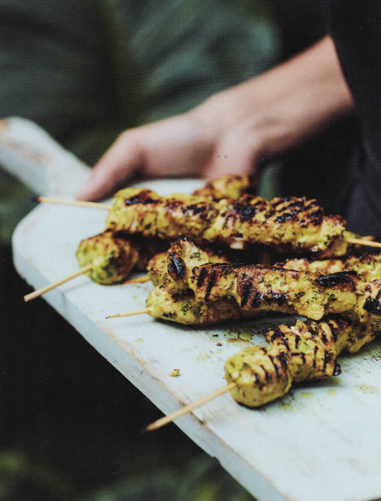Grilled chicken skewers on wooden sticks served on a white cutting board, held by a person.