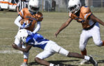 A football player in blue tackles a player in orange holding the ball, while another orange team player looks on during a game.