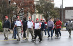 A group of people walk together on a city street during the day, some wearing matching red and white shirts and hats, with buildings and trees in the background.