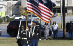 A group of uniformed individuals marching outdoors, carrying the American flag and another flag, with cars and people in the background.