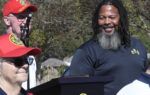 Three people at an outdoor event, two wearing red hats, one speaking at a podium, with trees and blue sky in the background.