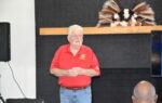 older man in a red shirt stands in front of a black fireplace with a decorative arrangement on the mantle.
