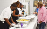 People serving food from trays at a buffet line, wearing gloves and aprons, while others wait to receive their meals indoors.