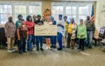 A group of people stands indoors holding a large ceremonial check, smiling and posing for a group photo in front of large windows.