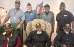 Six men pose together indoors, some seated and some standing, with one in uniform. An American flag is displayed in the background.