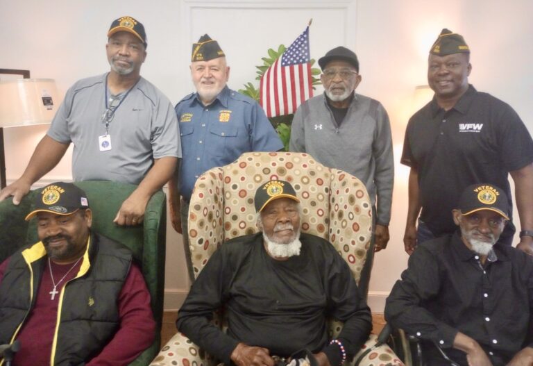Six men pose together indoors, some seated and some standing, with one in uniform. An American flag is displayed in the background.