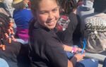 Smiling girl sitting in stadium bleachers among a crowd, wearing a black shirt and holding a blue object, with a football field in the background.