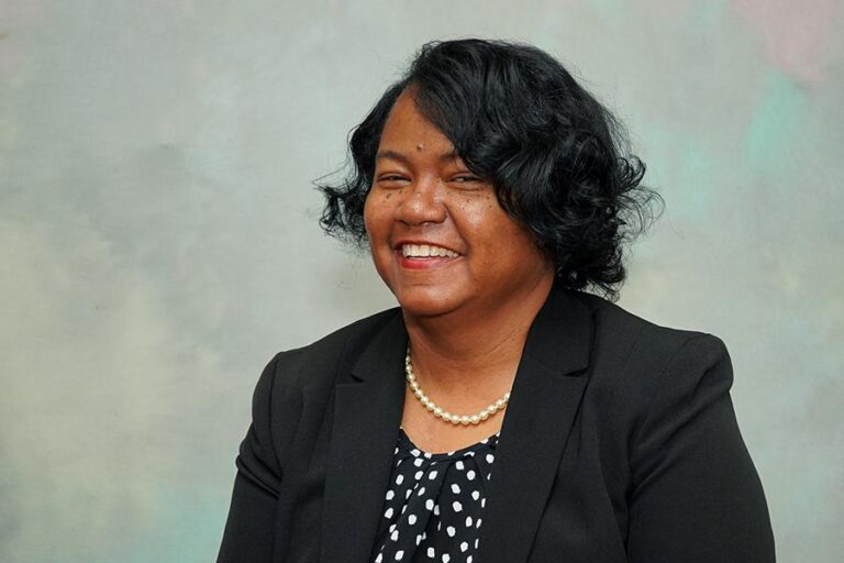 Smiling woman with black curly hair, wearing a black blazer, polka dot shirt, and pearl necklace, poses against a plain background.