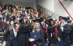 Graduates in caps and gowns celebrate at a graduation ceremony, smiling and holding diplomas, with a crowd of people seated in the background.