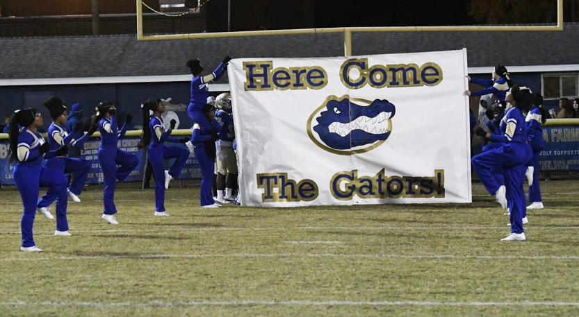 Cheerleaders hold a banner reading "Here Come The Gators" with a blue alligator logo on a football field at night.