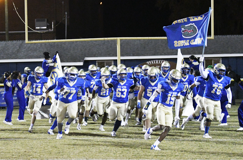 A football team in blue and gold uniforms runs onto the field at night, with one player waving a blue team flag.